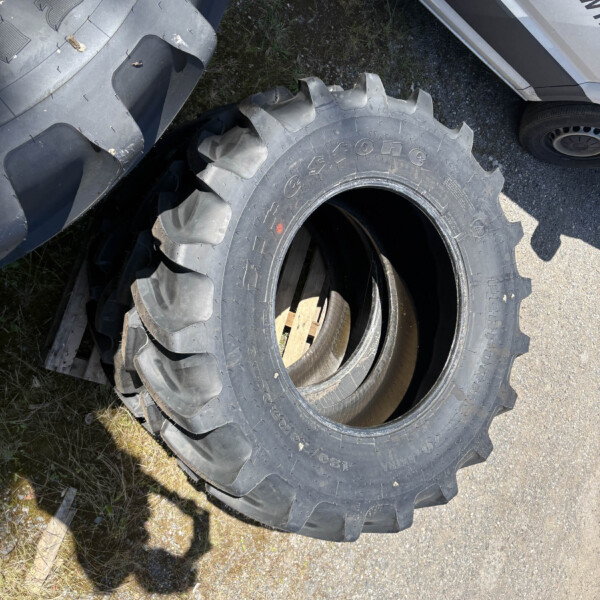Large rugged tractor or industrial tire with deep treads leaning against a stack of tires on a gravel surface nearby a white vehicle.