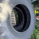 Large rugged tractor tire with deep treads lying on the ground beside a wooden pallet and green plants, outdoors.