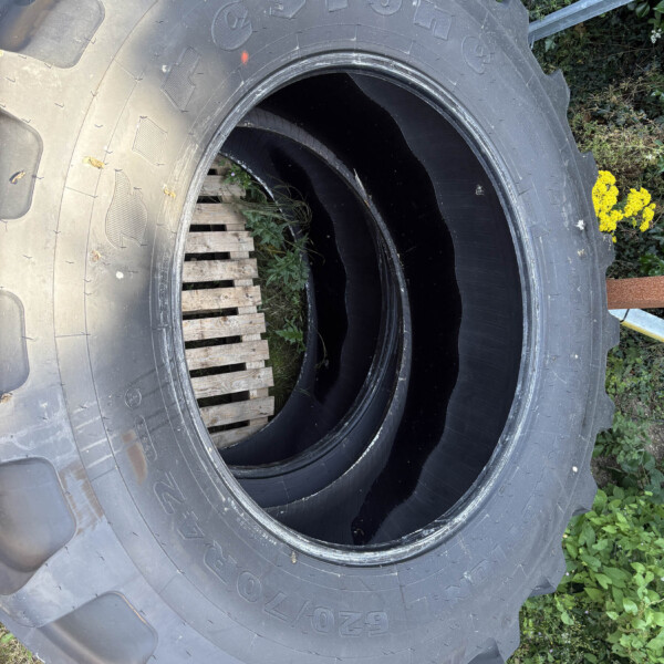 Large rugged tractor tire with deep treads lying on the ground beside a wooden pallet and green plants, outdoors.