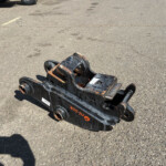 Rusty skid-steer attachment lying on an empty asphalt parking lot with faint yellow parking lines in the background.