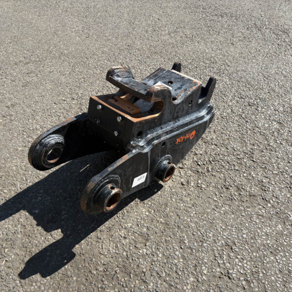 Rusty hydraulic breaker attachment resting on asphalt, showing bolts and orange paint.