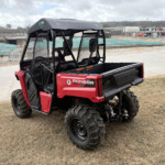 Red Honda Pioneer 520 side-by-side UTV parked on dry grass with an industrial worksite in the background.