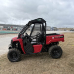 Red Polaris Pioneer 520 side-by-side utility vehicle with protective mesh cab parked on dry grass at an outdoor work yard, with trucks and buildings in the background under a cloudy sky.