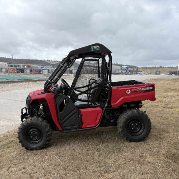 Red Polaris Pioneer 520 side-by-side utility vehicle with protective mesh cab parked on dry grass at an outdoor work yard, with trucks and buildings in the background under a cloudy sky.