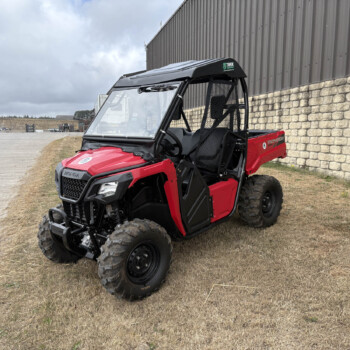 Red Honda Pioneer UTV parked on dry grass beside a stone wall and metal building, in an industrial yard setting.