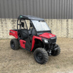Red Honda Pioneer utility side-by-side parked on dry grass beside a stone wall and metal siding.