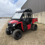Red Honda Pioneer side-by-side UTV parked on dry grass and dirt beside a brick wall and metal building in an industrial yard.