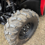 Close-up of a rugged off-road tire and suspension on a black vehicle, with dirt on the tire and a red body panel nearby.