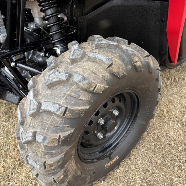 Close-up of a rugged off-road tire and suspension on a black vehicle, with dirt on the tire and a red body panel nearby.