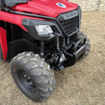 Close-up of a red Honda off-road vehicle front end with rugged tires and black grille, parked on dry grass by a stone wall.