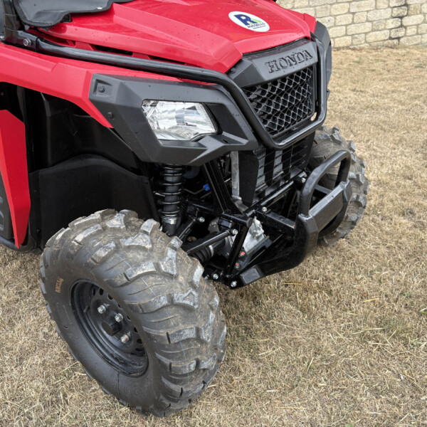 Close-up of a red Honda off-road vehicle front end with rugged tires and black grille, parked on dry grass by a stone wall.