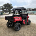 Rear view of a red Honda Pioneer 520 side-by-side utility vehicle parked on dry grass near an industrial yard.