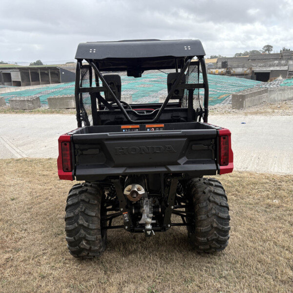 Rear view of a red Honda utility ATV with large off-road tires, parked on dry grass; damaged construction site or quarry buildings in the background under a cloudy sky.