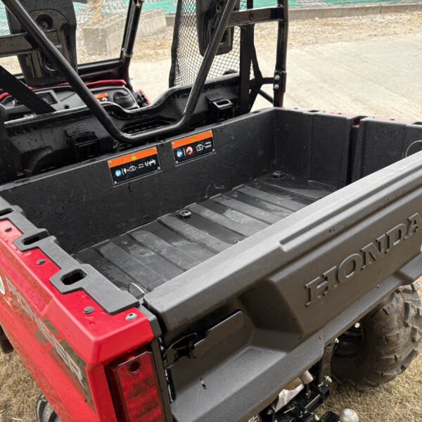 Red off-road utility vehicle seen from behind with open cargo bed and warning labels on tailgate, wet surface speckled with raindrops, tire tread visible.