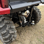 Rear-side view of a red off-road UTV showing large knobby tires, suspension, and a steel hitch on dry grass.