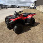 Red ATV parked on dry grass in an industrial yard with buildings and trucks in the background.