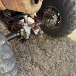 Rear view of a dusty tractor undercarriage showing a large treaded wheel and exposed suspension components on dry grass ground.
