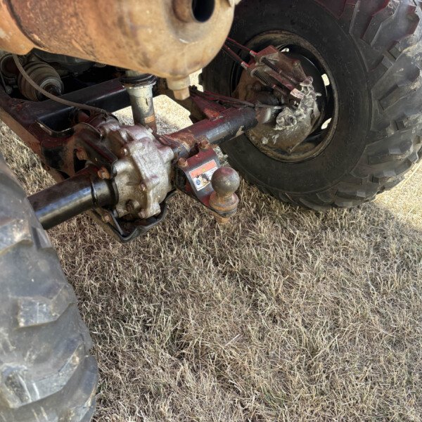 Rear view of a dusty tractor undercarriage showing a large treaded wheel and exposed suspension components on dry grass ground.