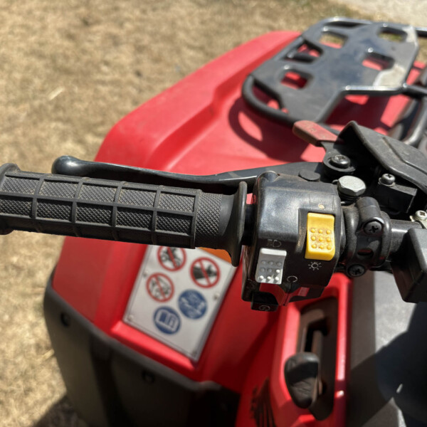 Close-up of a black textured handlebar grip and throttle on a red vehicle, with nearby controls and a warning label visible on the body.