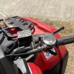 Close-up of red ATV handlebar area with a brake fluid reservoir cap and textured grip visible.