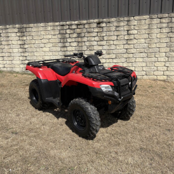 Red ATV with black rack sits on dry grass in front of a stone masonry wall and metal fence.