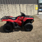 Red ATV parked on dry grass beside a textured stone wall, with industrial buildings in the background.