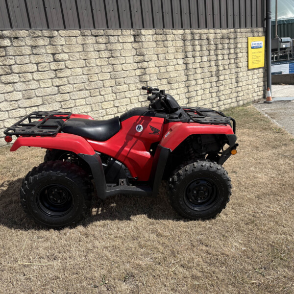 Red ATV parked on dry grass beside a textured stone wall, with industrial buildings in the background.