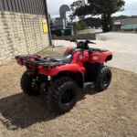 Red Honda ATV parked on dry grass beside a stone wall, with industrial buildings and a concrete driveway in the background.