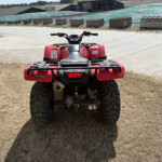 Red ATV viewed from behind in a dry grassy area with an industrial complex in the distance; large knobby tires and rear rack visible.