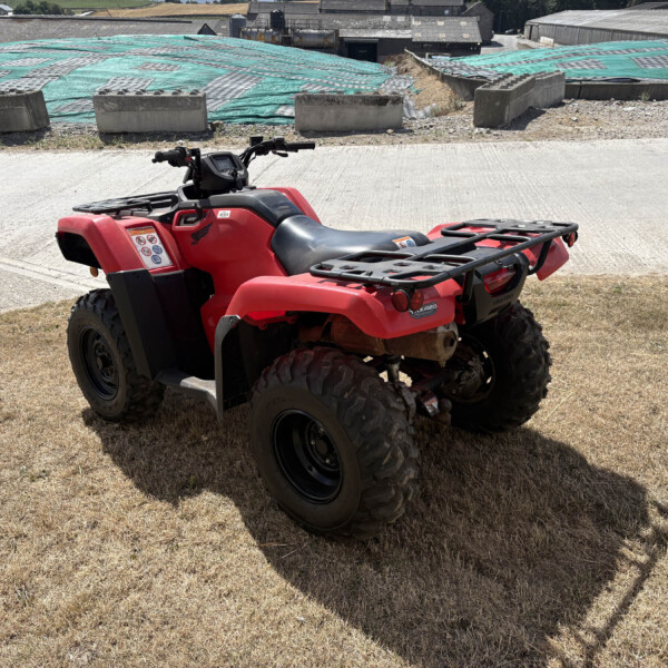 Red ATV parked on dry grass beside a concrete path, with a construction area and green tarps in the background.