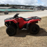 Red all-terrain vehicle (ATV) parked on dry grass with an industrial site in the background and clear partly cloudy sky.