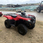Red ATV and farming equipment parked on a muddy field with a tractor in the background and farm buildings nearby.