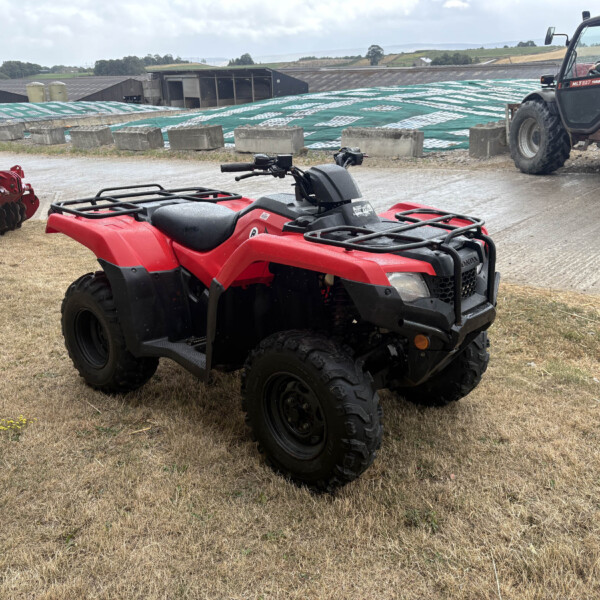 Red ATV and farming equipment parked on a muddy field with a tractor in the background and farm buildings nearby.