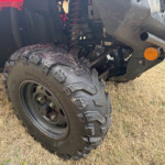 Close-up of a muddy off-road vehicle wheel and suspension with an orange bumper marker light visible at the front corner.
