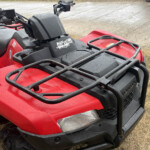 Close-up of a red ATV front with a black front rack and wet surface after rain, showing the headlight and handlebars edgeek