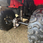 Underside view of a red ATV showing the rear suspension, coil spring, and large muddy tires with rusty metal components.