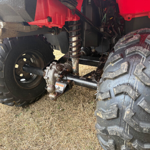 Underside view of a red ATV showing the rear suspension, coil spring, and large muddy tires with rusty metal components.