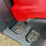 Close-up of a red car body panel meeting a black textured step bumper, with a metal strap and dirt specks visible on the surface.