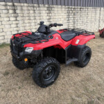 Red ATV with black racks parked on dry grass beside a stone wall, front view showing rugged tires and handlebar area.