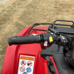 Close-up of red ATV handlebars with the yellow brake lever and warning label, outdoors on dry grass/dirt area. (informative)
