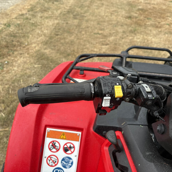 Close-up of red ATV handlebars with the yellow brake lever and warning label, outdoors on dry grass/dirt area. (informative)