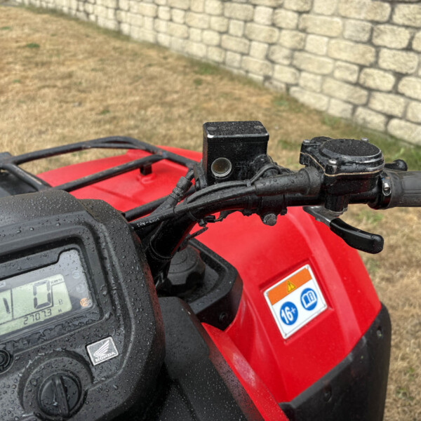 Close-up of a red motorcycle/ATV cockpit with a rain-speckled display and handlebars, stone wall in the background.
