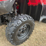 Close-up of an off-road vehicle's front wheel and suspension with dirt and an orange side marker light nearby.