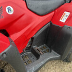 Red Honda ATV with black step rails, logo and a circular Russell sticker on the body, sitting on dry grass; rain droplets on surface.
