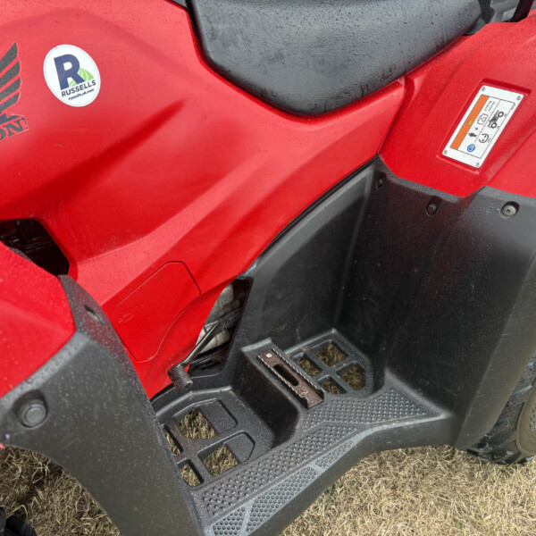 Red Honda ATV with black step rails, logo and a circular Russell sticker on the body, sitting on dry grass; rain droplets on surface.