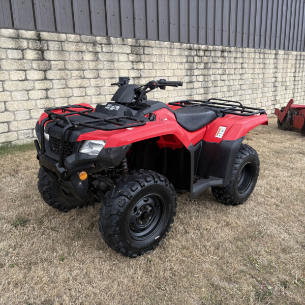 Red ATV parked on dry grass beside a stone wall and corrugated metal building.