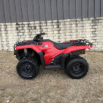 Red ATV parked on dry grass in front of a stone wall and a wooden fence.