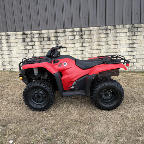 Red ATV parked on dry grass in front of a stone wall and a wooden fence.