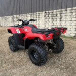 Rear three-quarter view of a red Honda ATV with black racks parked on dry grass beside a stone retaining wall and corrugated metal building.