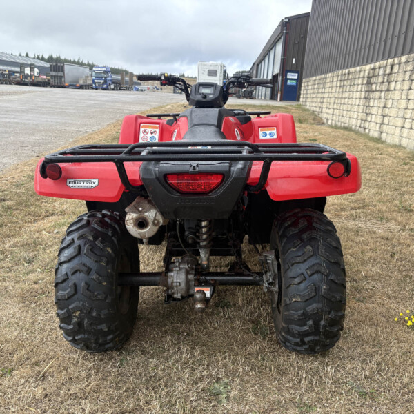 Rear view of a red Honda Fourtrax ATV on a dry grassy lot with an industrial yard in the background
